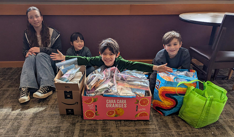 A group of four children sit on the floor of a room with a carpeted floor and wooden furniture. They are behind several boxes and reusable bags filled with assembled care packages in plastic bags. The child in the center, wearing a green long-sleeve shirt, has arms outstretched with a big smile. The child on the left, wearing glasses and a dark hoodie, sits with legs crossed, smiling gently. The other two children, one in a dark gray shirt and the other in a blue shirt, also smile at the camera. The care packages appear to contain snacks and hygiene items, and the setting suggests a volunteer effort.