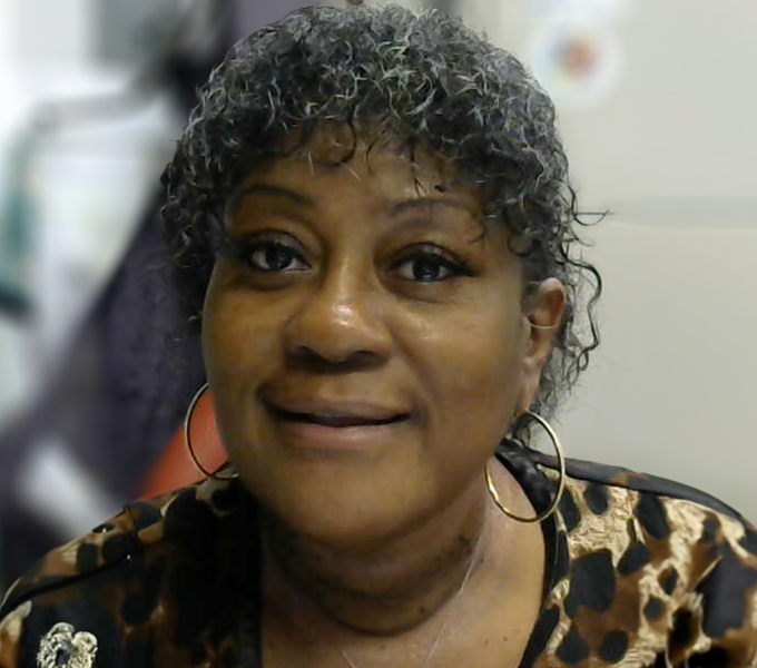 A woman with short, curly black hair is smiling at the camera. She is wearing large gold hoop earrings and a black and brown leopard-print top with a brooch on the left side. The background includes an office setting with a filing cabinet. A circular sticker with a colorful design is visible on the cabinet.