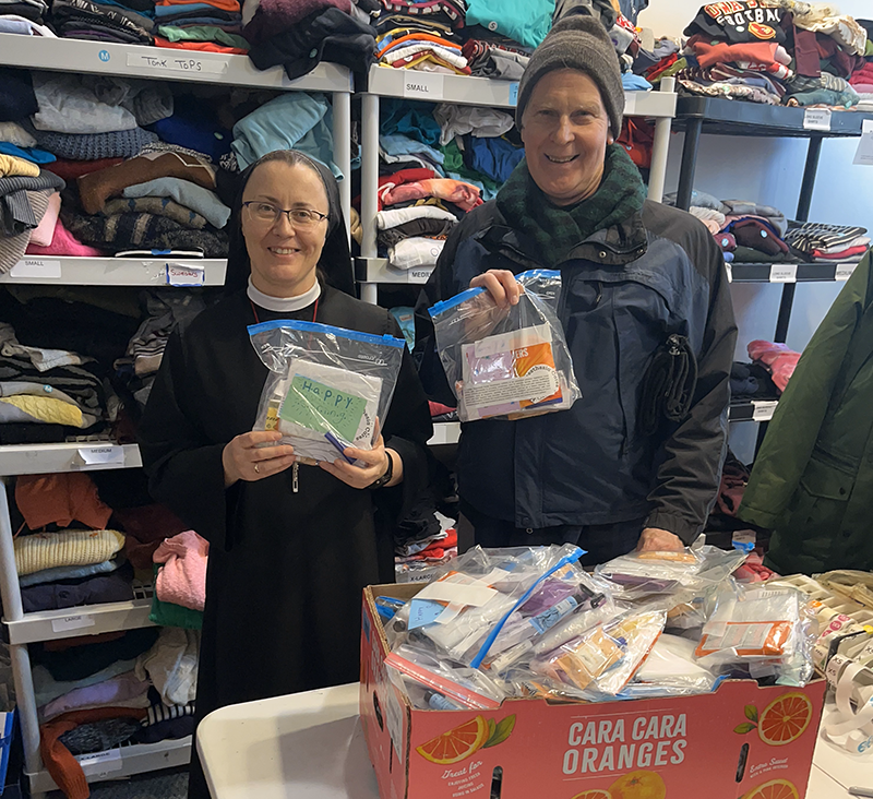 A nun and a man stand in a room filled with shelves stacked with folded clothing. They are both smiling and holding clear plastic bags containing care package items, such as snacks and hygiene products. The nun’s bag has a handwritten note that reads “HAPPY.” In front of them is a large cardboard box labeled “Cara Cara Oranges,” filled with more care packages. The setting appears to be a donation or volunteer center, where supplies are being prepared for distribution.