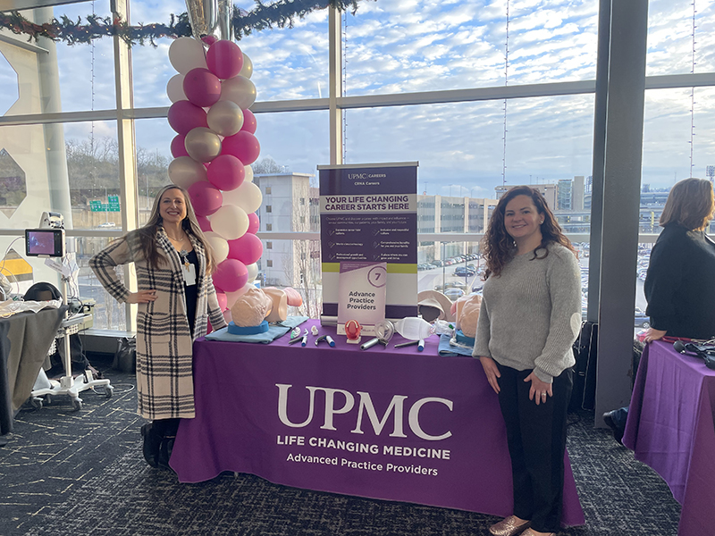 Two women stand on either side of a table covered with a purple tablecloth that reads "UPMC Life Changing Medicine, Advanced Practice Providers." Behind the table is a display with a sign that says, "Your life-changing career starts here" and promotes opportunities for advanced practice providers. The table features medical training tools, including anatomical models and medical instruments. A balloon column in pink and white stands to the left, with large windows in the background revealing a parking lot and a partly cloudy sky.