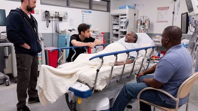 Image from Season 1, Episode 8 of "The Pitt": Dr. Langdon and Dr. Robby in an ER exam room with patient Willie lying on a hospital bed while his son sites nearby