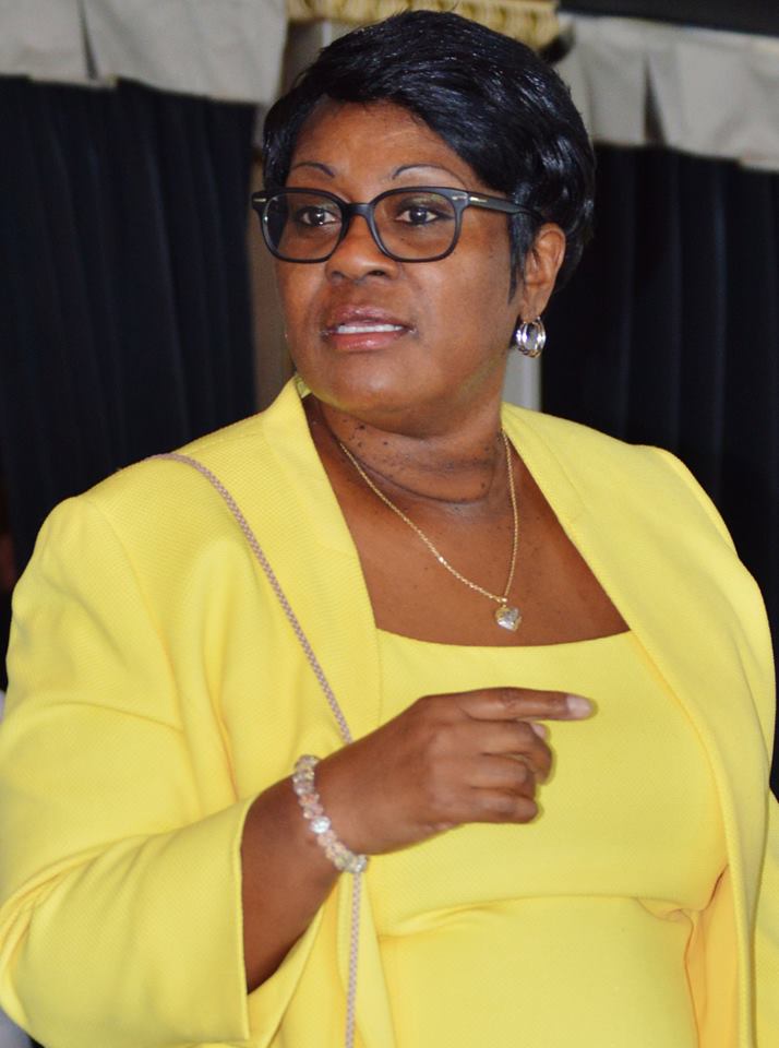 A woman with short black hair and glasses is wearing a bright yellow blazer over a matching dress. She is accessorized with a gold necklace with a heart-shaped pendant, pearl earrings, a bracelet, and a beige shoulder bag. She appears to be speaking or gesturing with her right hand, with a confident expression on her face. The background features dark curtains and a partially visible white canopy.