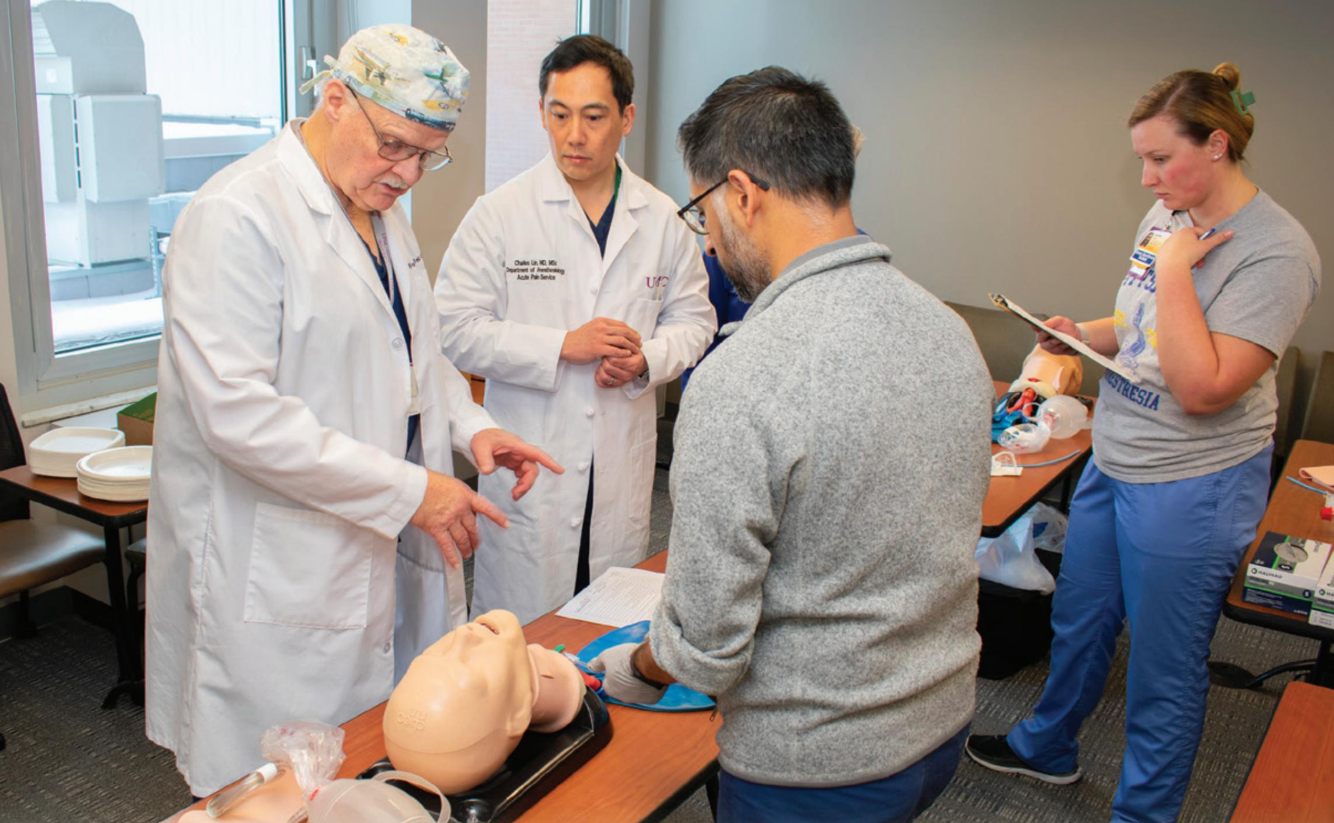 Alt text: A group of medical professionals participate in a hands-on training session in a classroom setting. A clinician wearing a white lab coat and a surgical cap gestures while explaining a procedure to a man in a gray sweater, who is practicing on a medical mannequin head. Another clinician in a white coat observes, while a woman in scrubs takes notes on a clipboard. Medical training equipment and mannequins are visible on the tables.