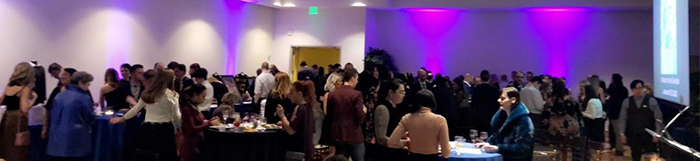 "A panorama view of the dinner event, people sitting and standing at tables in formal wear"