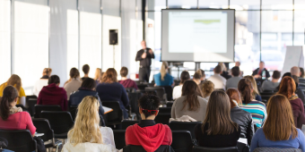 Photo of people at a lecture, looking at the speaker presenting with a PowerPoint