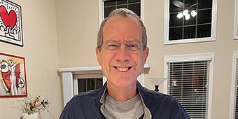 A smiling older man with short gray hair, glasses, and light skin stands in a warmly lit living room. He is wearing a navy blue pullover with a Detroit Tigers logo and a gray T-shirt underneath. His hands rest on a white countertop as he looks at the camera. The background features a beige couch, a patterned rug, framed artwork on the walls, and large windows with white blinds.